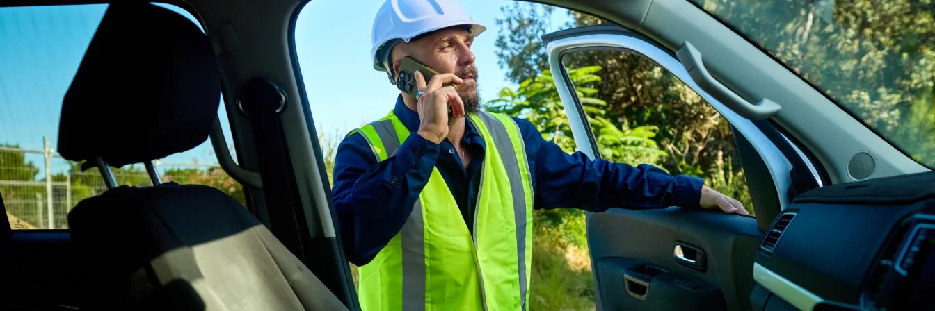 Field services worker in blue hard hat and high-visibility vest talking on phone while sitting in vehicle.