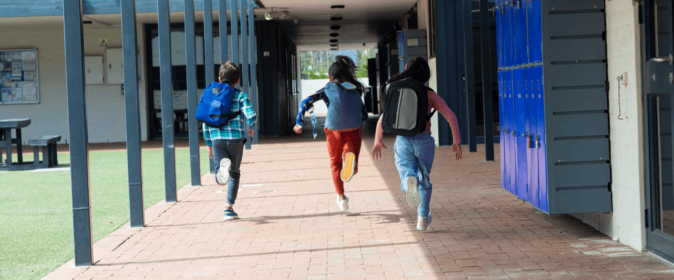  Three schoolchildren sprint past lockers and down an outdoor hallway.