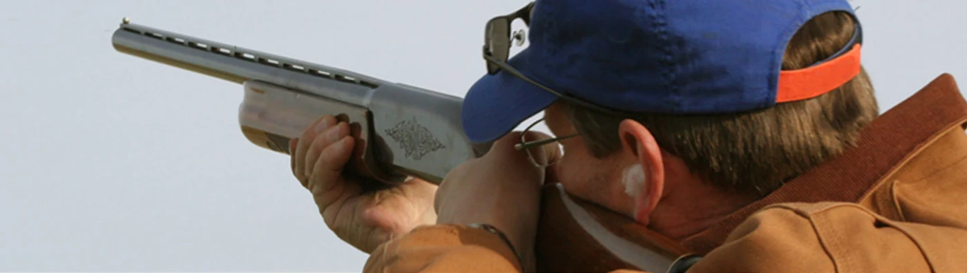 Man aiming a shotgun while wearing a blue cap, ear protection, and shooting glasses.