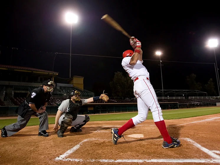Baseball batter swings under stadium lights, with catcher and umpire watching closely.