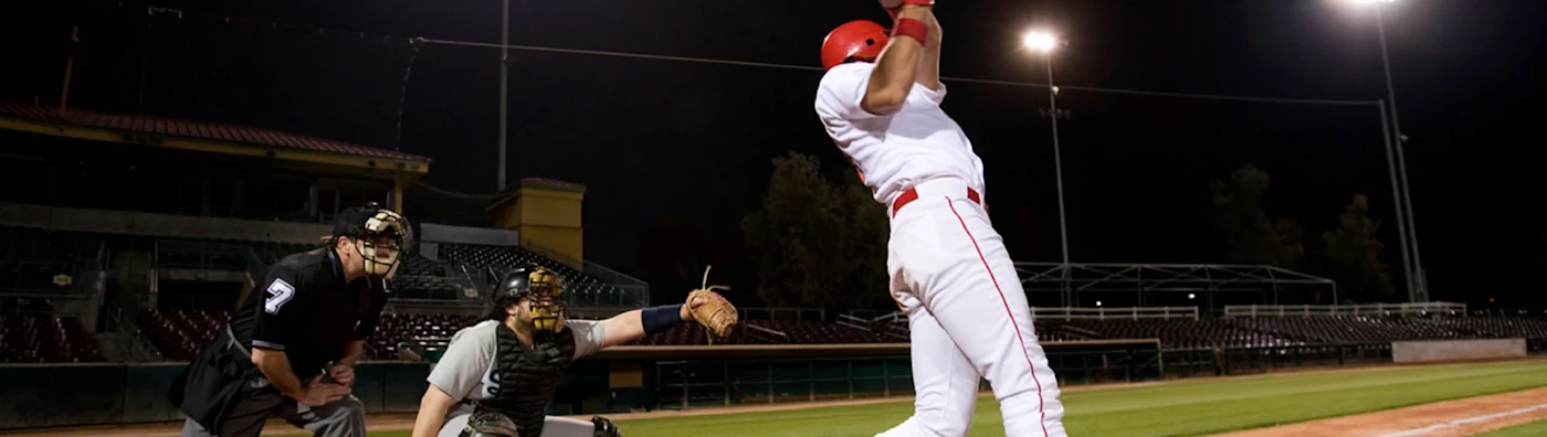 Baseball batter swings under stadium lights, with catcher and umpire watching closely.