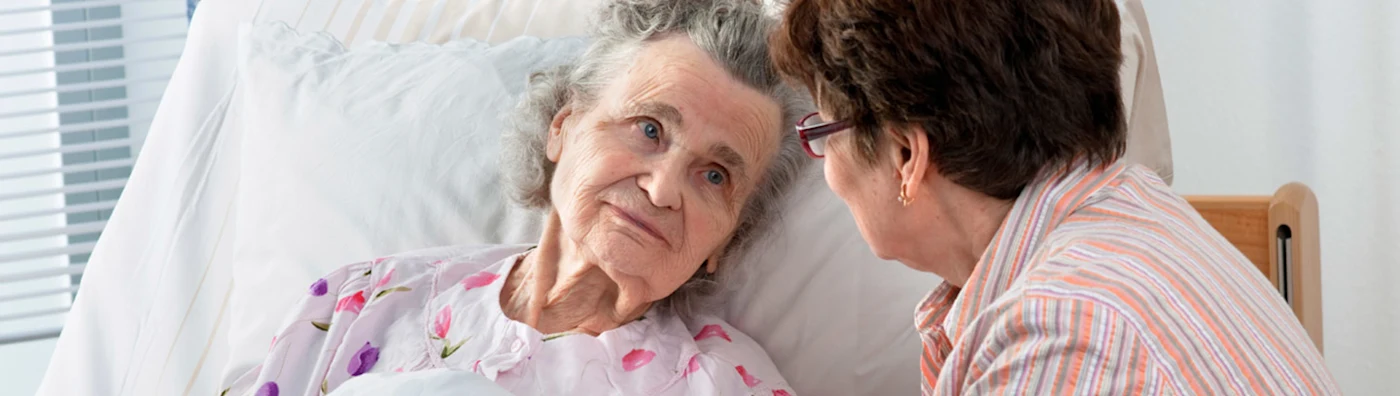Cose up image of an elderly woman lying in bed looking at another woman sitting beside her.