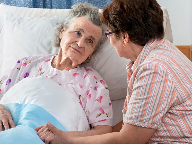 Cose up image of an elderly woman lying in bed looking at another woman sitting beside her.
