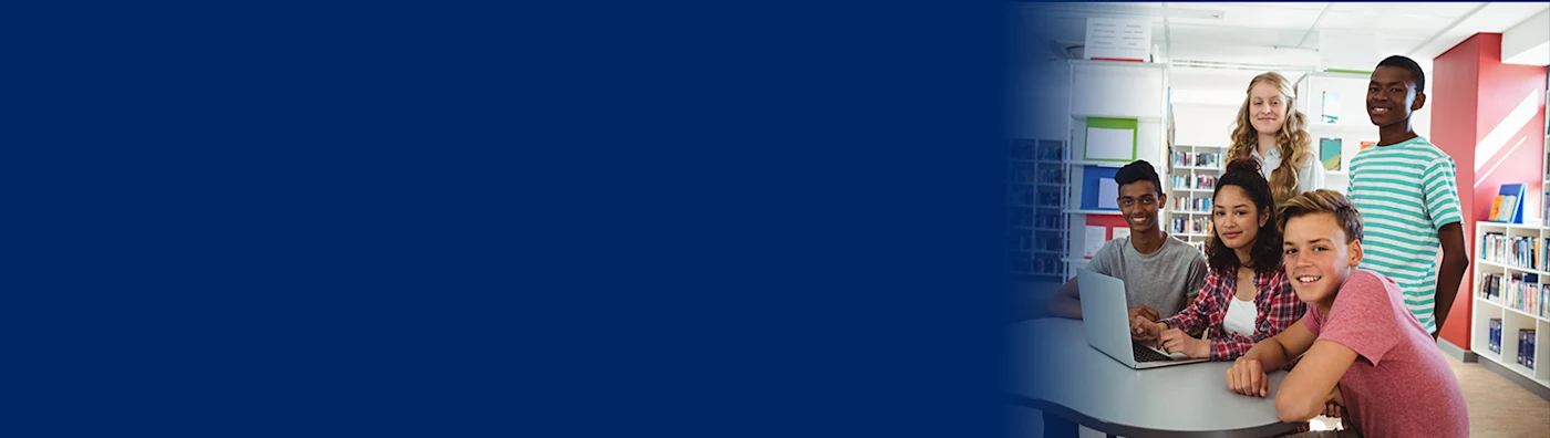 Group of young students smiling and gathered around a laptop in a school library. Left side of image is blurred.