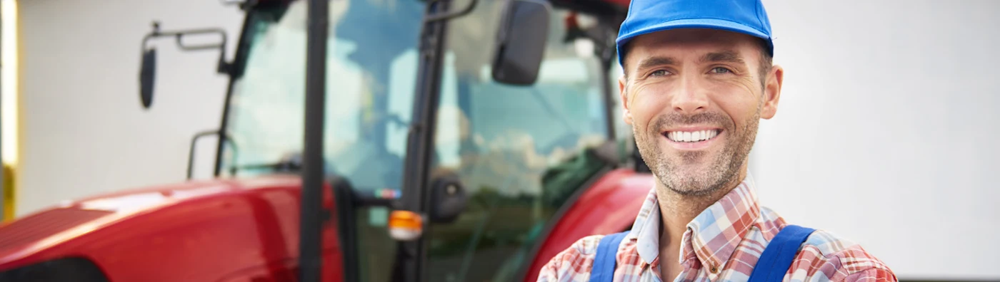 A male farmer stands outside in front of his red tractor parked in front of a barn.