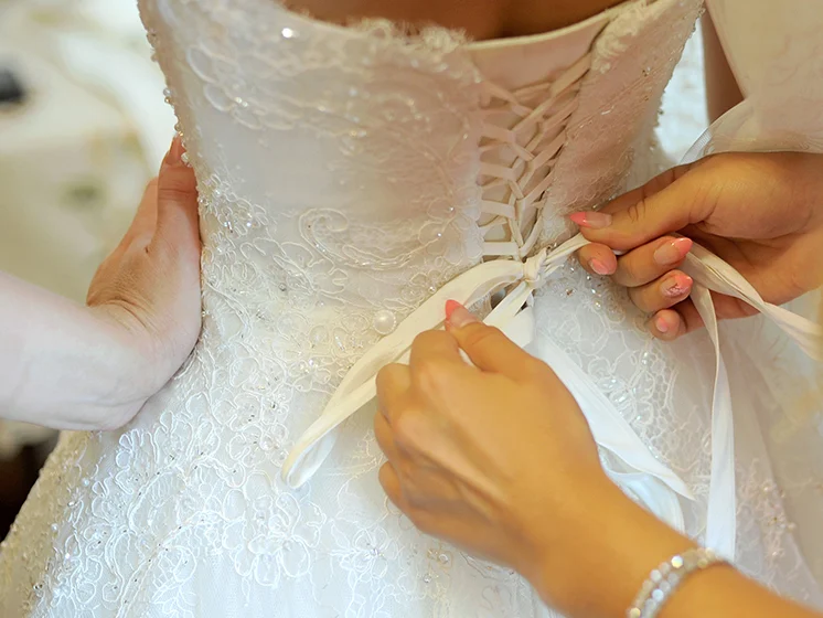 Close up image of hands tying the lace back of a detailed white wedding dress on a bride.
