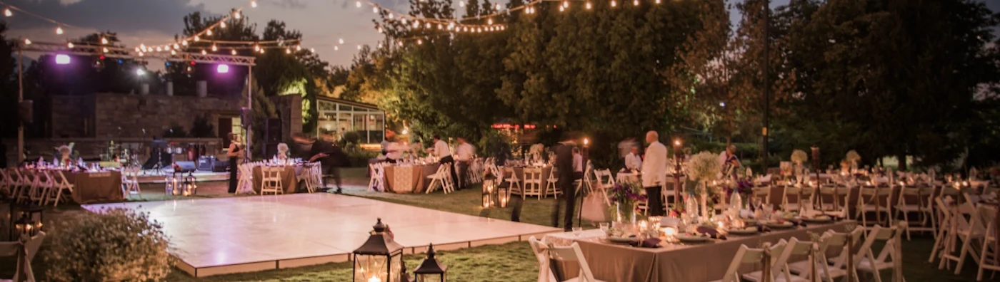 Outdoor wedding reception setup at dusk with string lights, dining tables, and dance floor.