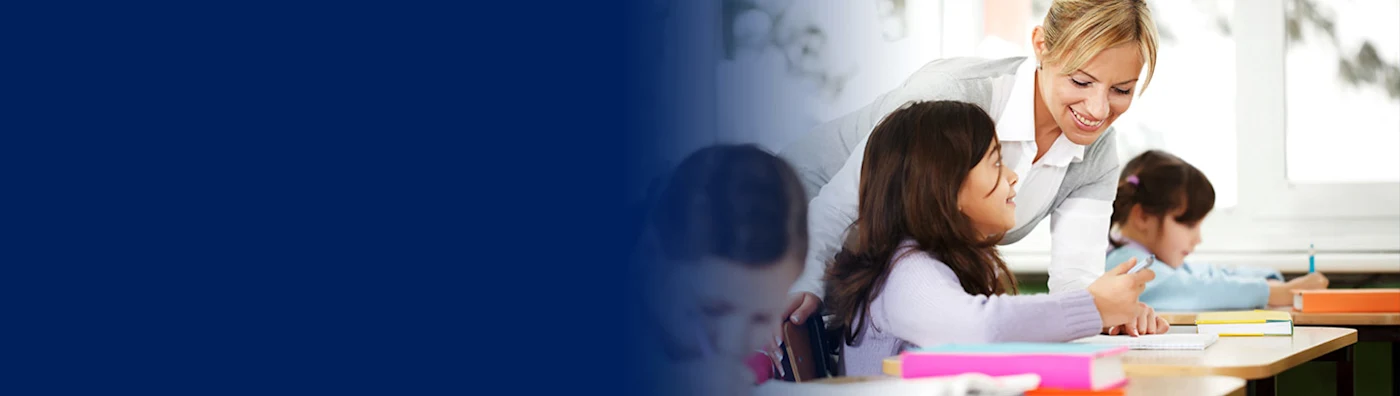 Smiling teacher helping a young student in a classroom with other children working nearby.