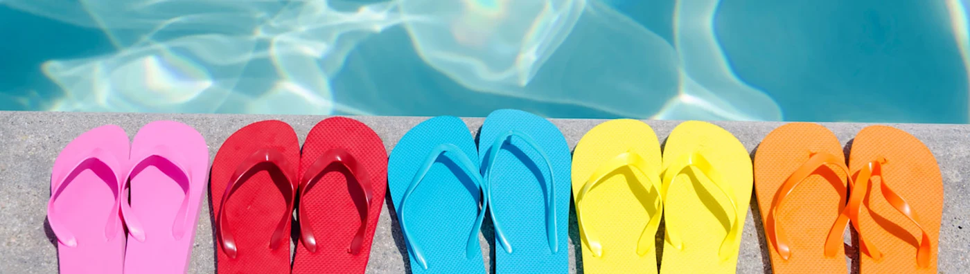 5 pairs of flip flops lined by a swimming pool on a sunny day.