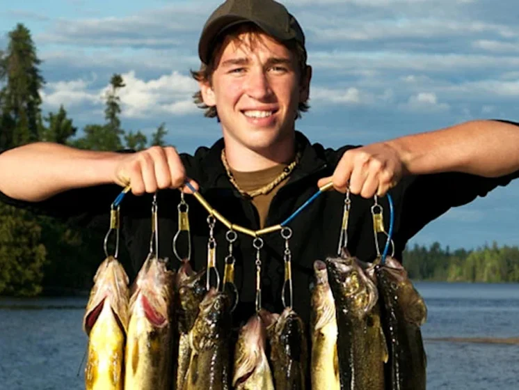 Young man proudly holds up his catch at a lakeside dock, with boats and forest in the background.