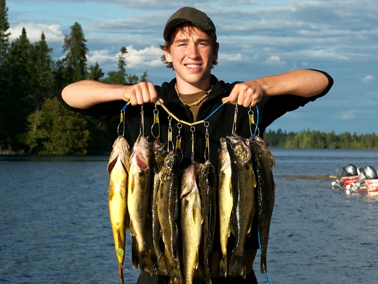Young man proudly holds up his catch at a lakeside dock, with boats and forest in the background.