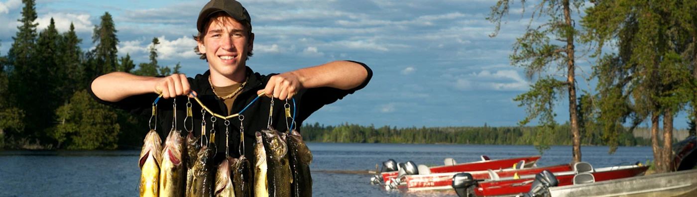 Young man proudly holds up his catch at a lakeside dock, with boats and forest in the background.
