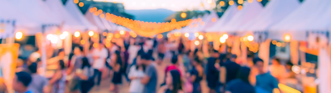 Blurred view of a lively outdoor market at dusk with string lights and white vendor tents.