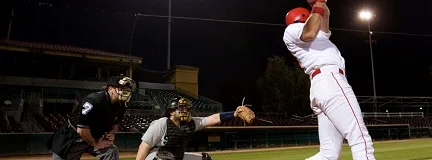 Baseball batter swings under stadium lights, with catcher and umpire watching closely.