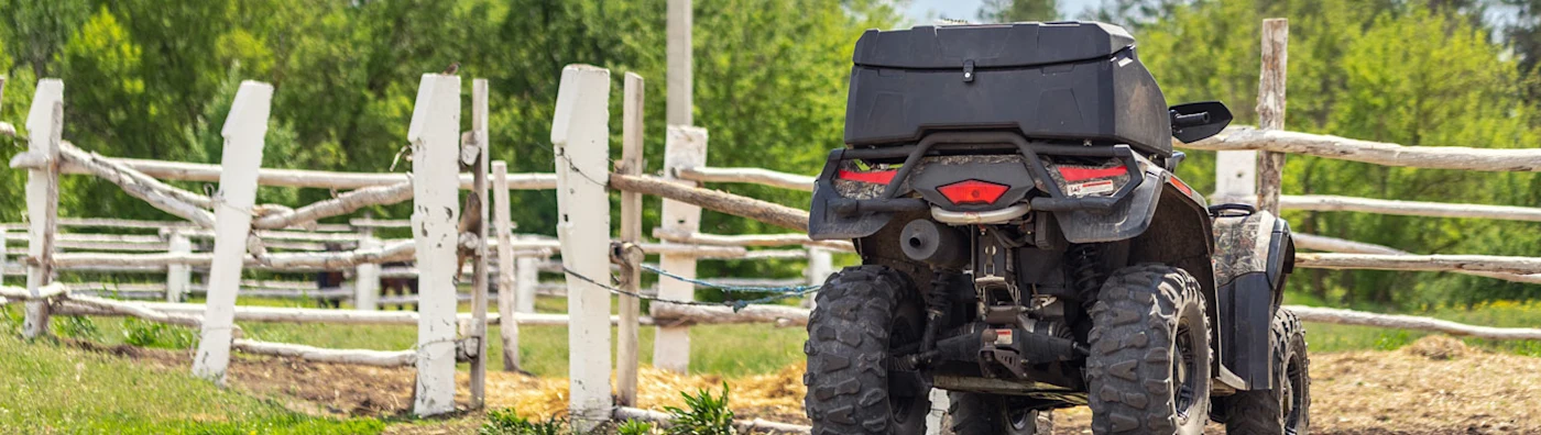 An ATV parked near a rustic wooden fence on a farm with trees in the background.