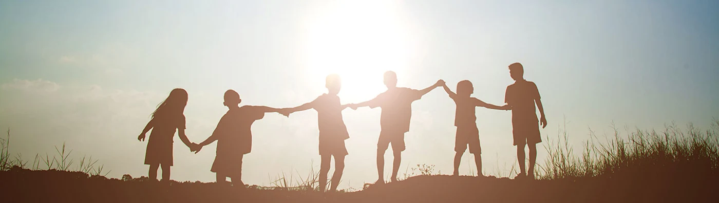 Silhouetted children holding hands in a line at sunset, standing on a grassy hill.