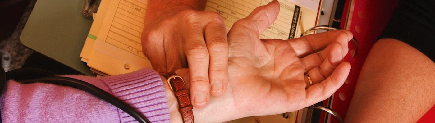 Person checks another's pulse by hand over a clipboard filled with note.