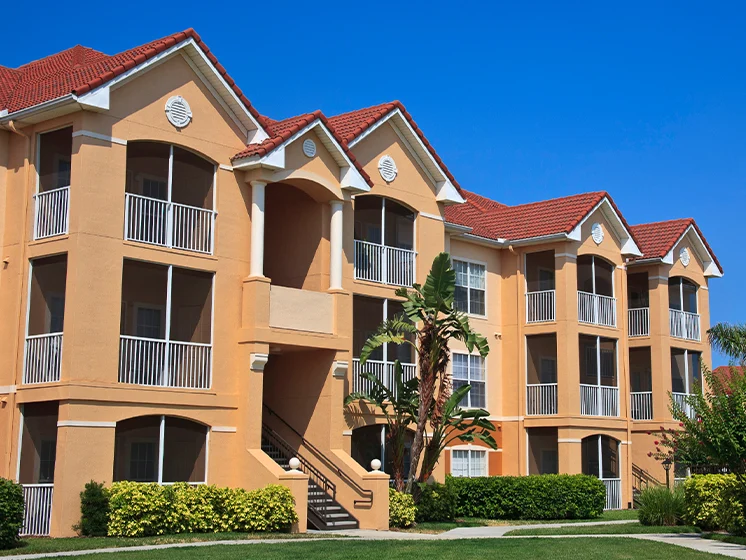 Modern condominium building with tan walls, red tile roof, and balconies, set against a clear blue sky.