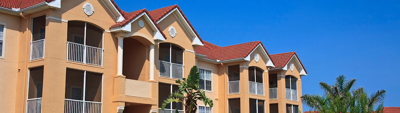 Modern condominium building with tan walls, red tile roof, and balconies, set against a clear blue sky.