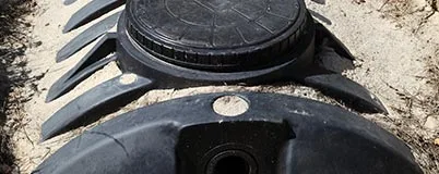 Close up of a black septic tank lid and riser partially buried in sand or soil.