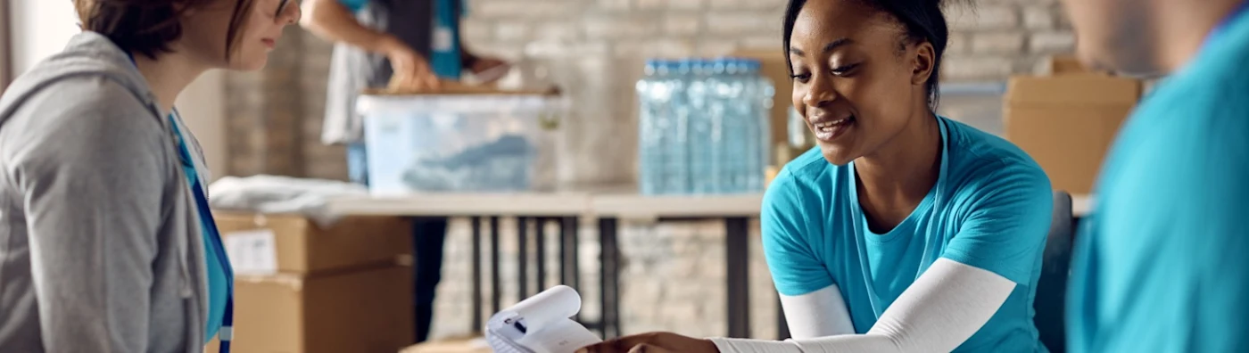 Volunteers review a checklist in brick building with volunteer in the background packing a box.
