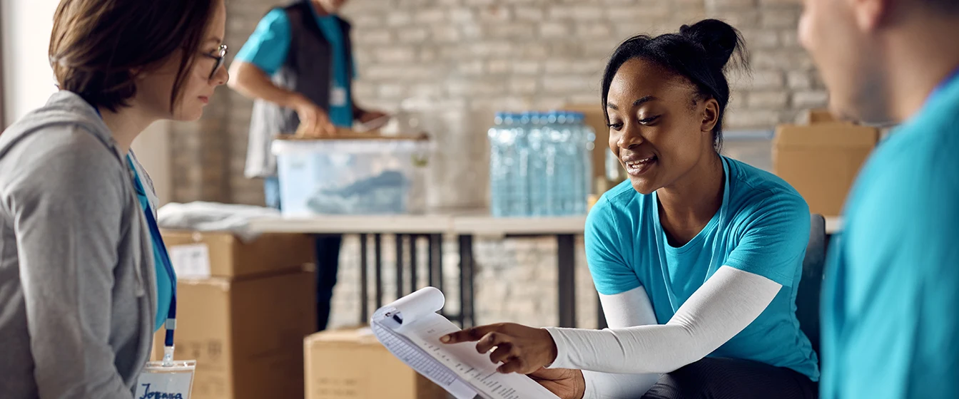 Volunteers review a checklist in brick building with volunteer in the background packing a box.
