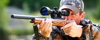 Man in camo gear aiming rifle with scope, outdoors in a wooded area during daylight.