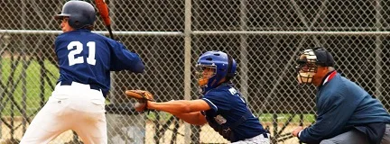 Baseball player at bat, with catcher and umpire ready behind home plate on a fenced field.