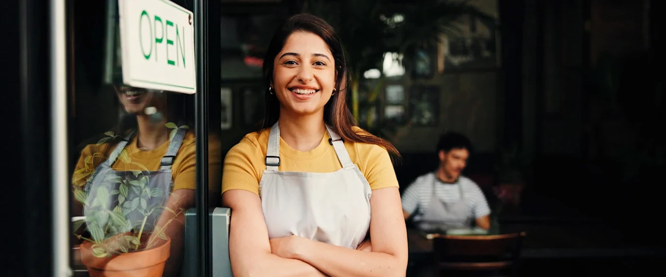 A smiling business owner stands next to her cafe’s open door