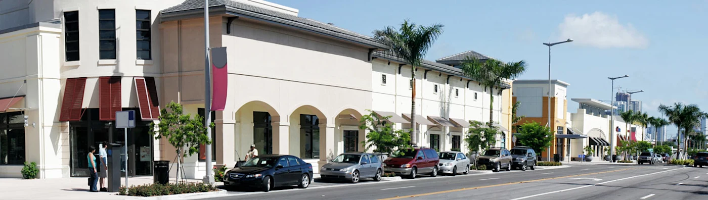 Modern commercial shopping plaza with palm trees, storefronts, and parked cars along the street.