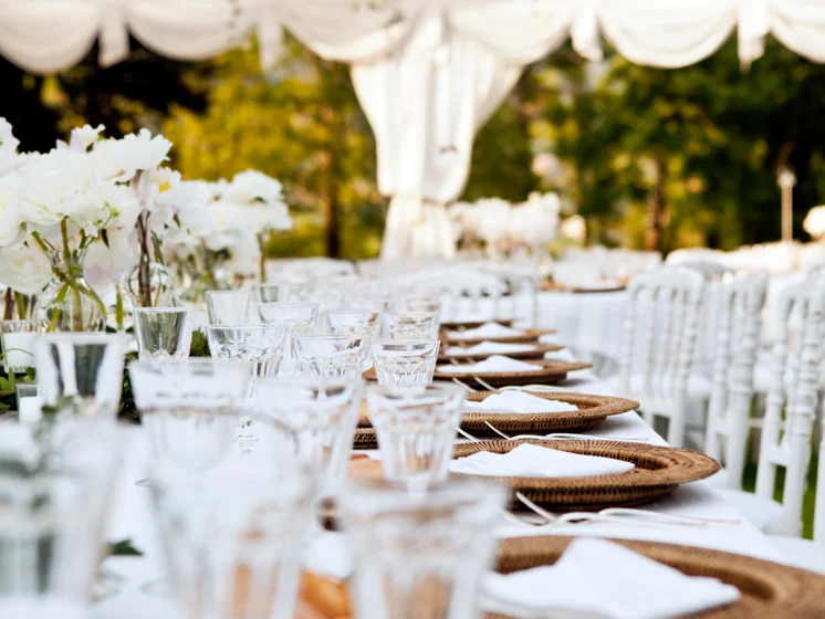 Elegant outdoor table setting with white chairs, woven chargers, glassware, and floral centerpieces.