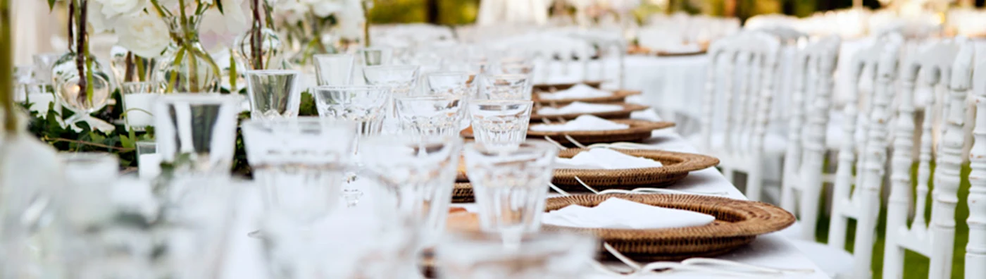 Elegant outdoor table setting with white chairs, woven chargers, glassware, and floral centerpieces.
