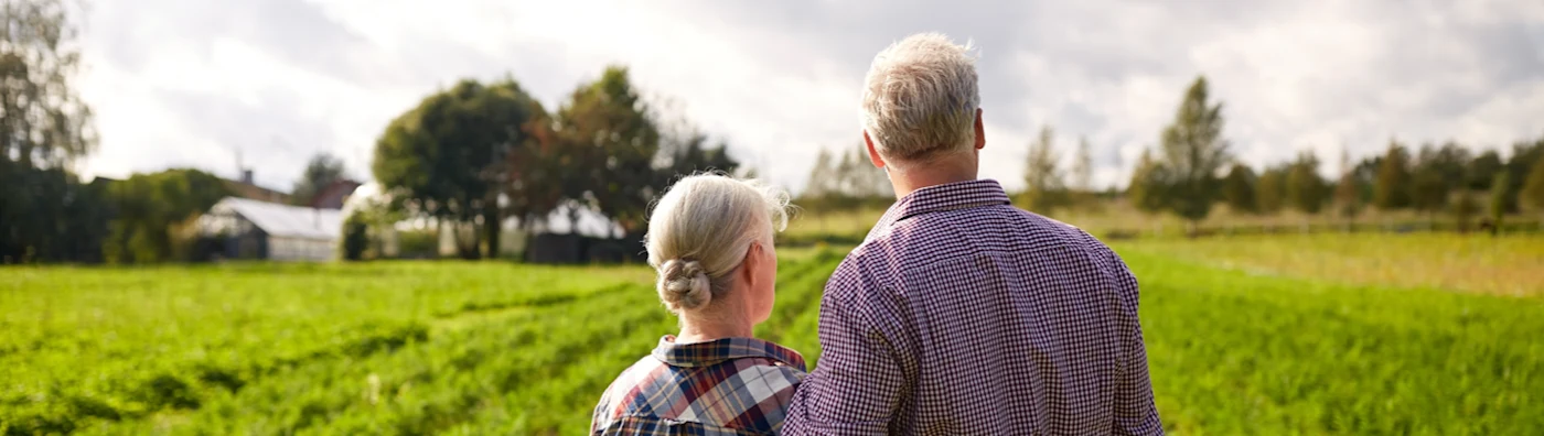 An older couple stands in their farm’s field among the crops with their arms wrapped around each other's waists.