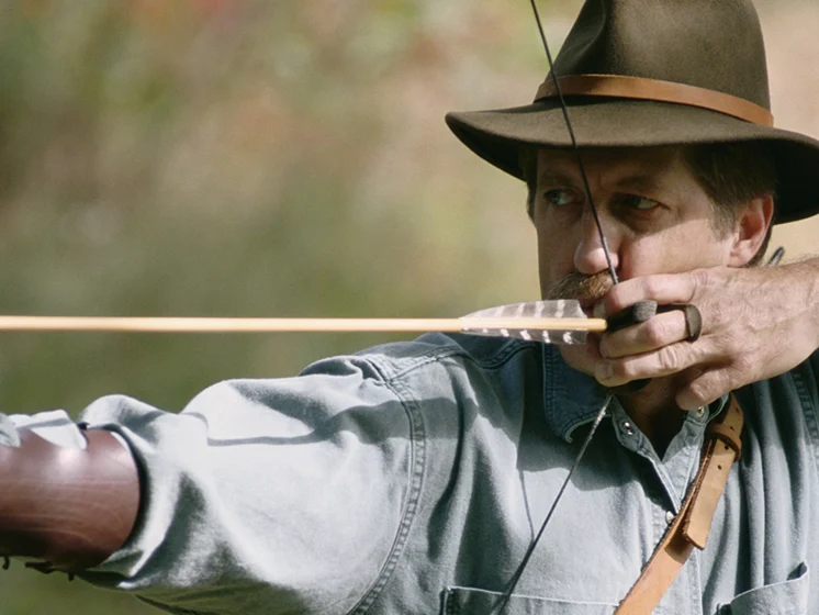 Man wearing a hat draws a bow, aiming an arrow in an outdoor setting.