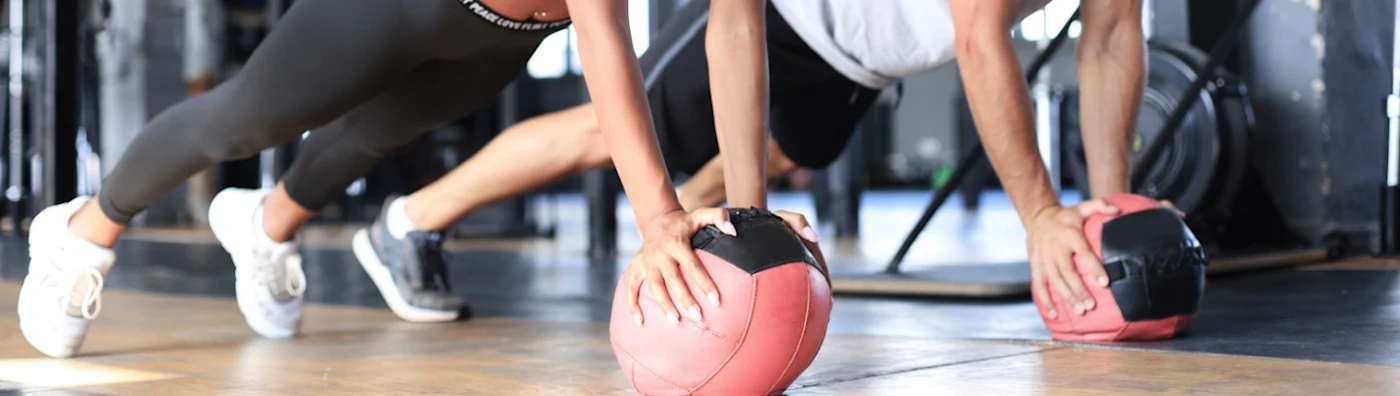 Young man and woman exercising with medicine ball in gym, looking ahead; gym equipment is blurred in the background.