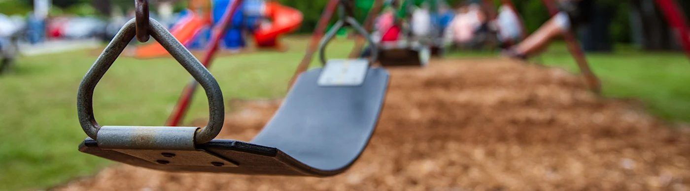 Close-up of an empty playground swing with blurred background of kids and colorful equipment.