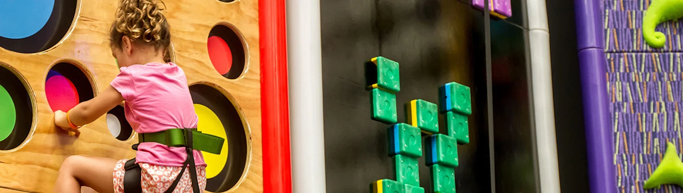 A child in a safety harness climbs a colorful indoor wall, part of a family entertainment center.