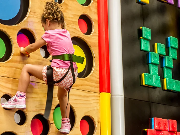 A child in a safety harness climbs a colorful indoor wall, part of a family entertainment center.