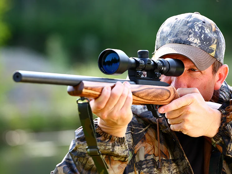 Man wearing camouflage aims a scoped rifle outdoors, focusing through the sight.