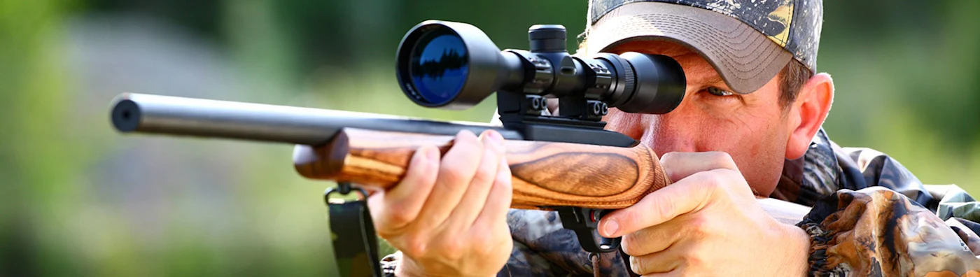 Man wearing camouflage aims a scoped rifle outdoors, focusing through the sight.