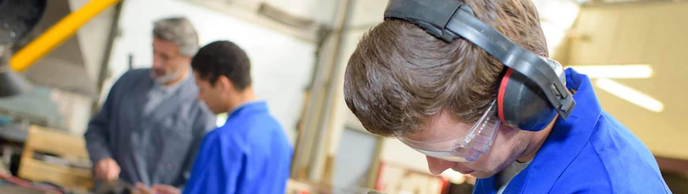 Man wearing safety gear focuses on a task in a workshop, with others working in the background.