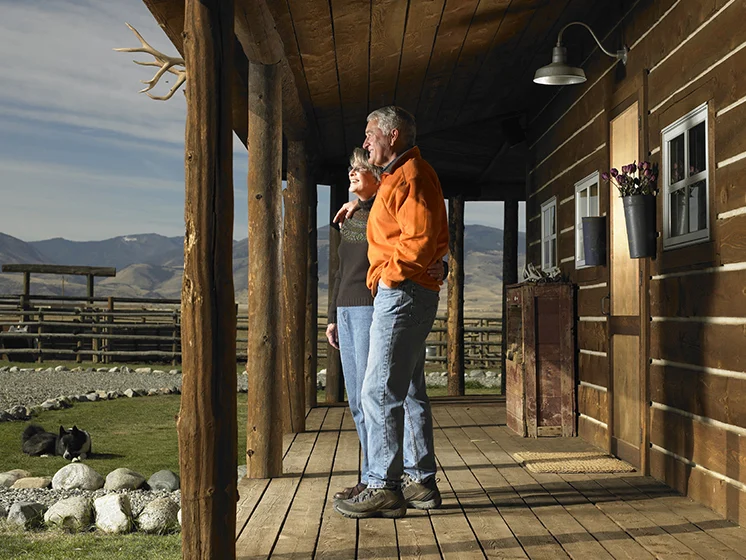 A couple stands on the porch of a rustic wooden lodge, enjoying a scenic mountain view.