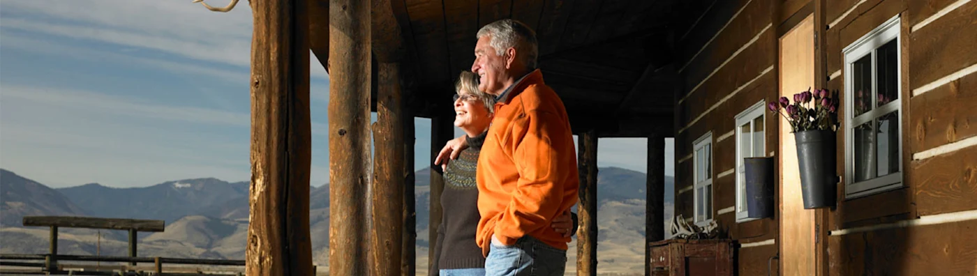 A couple stands on the porch of a rustic wooden lodge, enjoying a scenic mountain view.