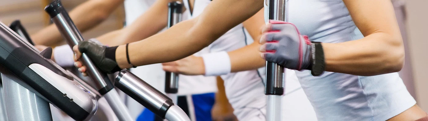 Close-up of people exercising on elliptical machines in a gym, wearing workout gloves.