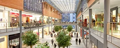 Spacious modern shopping mall with two levels, storefronts, indoor trees, and people walking around.