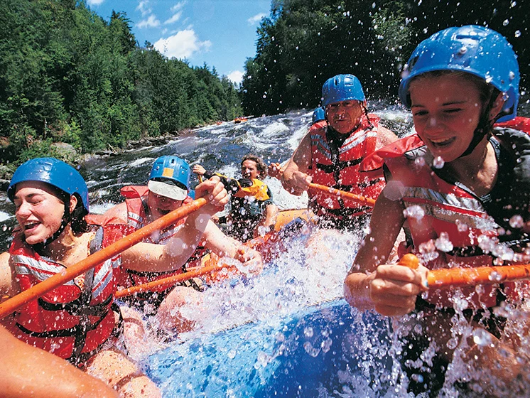 Close up image of a group of people wearing helmets and life vests whitewater rafting through a river.