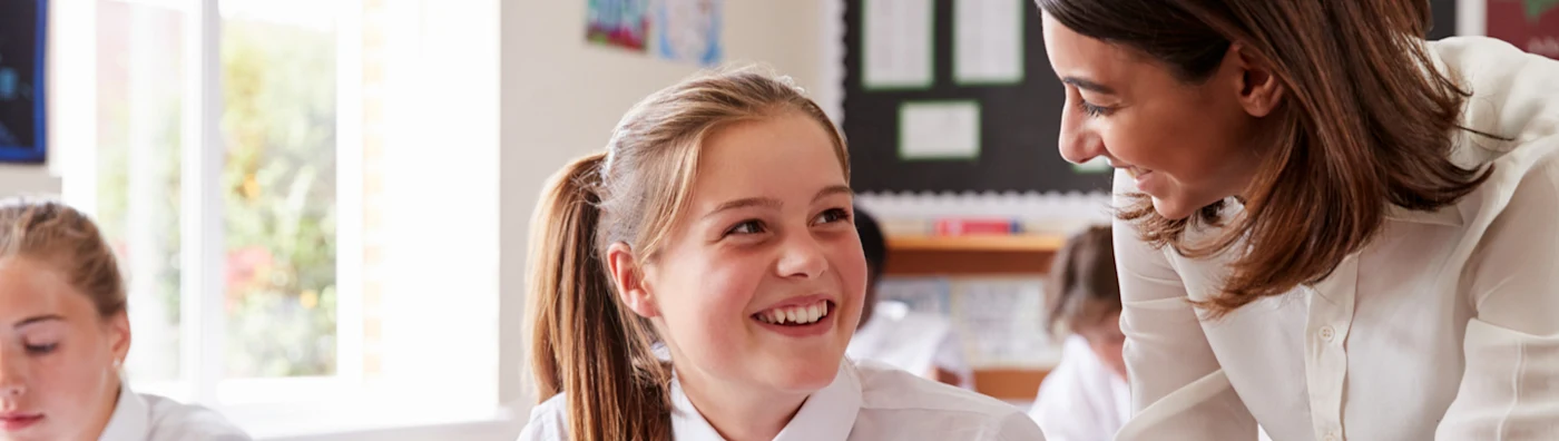 Teacher smiling at student in a bright classroom, both wearing white uniforms.
