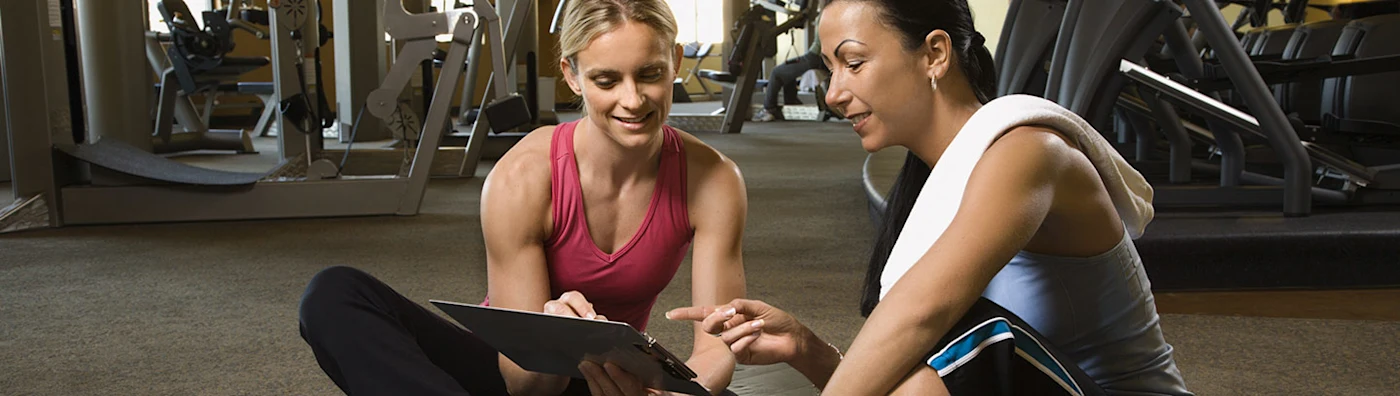Two women sitting on a gym floor smiling and reviewing a clipboard together near workout equipment.