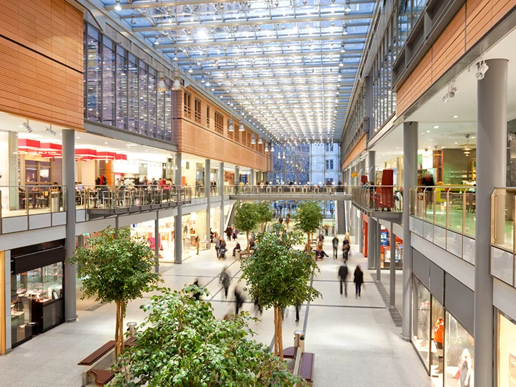 Spacious shopping mall with two levels, storefronts, indoor trees, and people walking around.