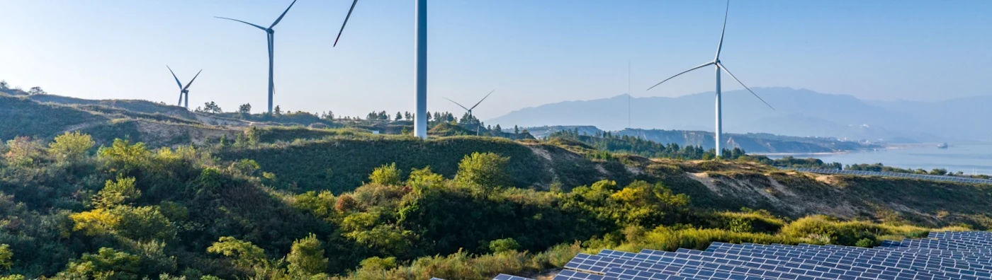 Solar panels and wind turbines on a hill with a view of mountains in the distance.  
